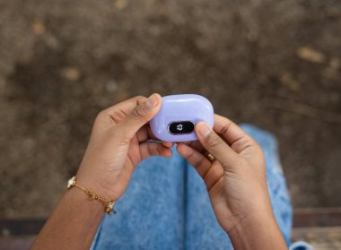 Mor kablosuz, şık bir kulaklık taşıyan eller şarj kutusu, pil seviyesi için bir LED göstergesi sergiliyor, sakin bir açık hava parkındaki bankta dinleniyor.