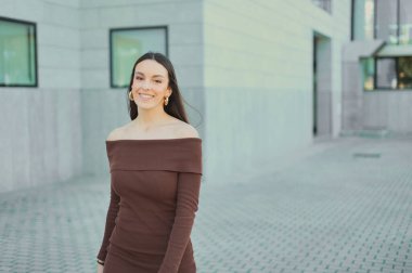 Esmer bir kadın moka ya da kahverengi, omuzları açık bir elbise giyiyor. Arka planı bulanık modern bir binanın yakınında gülümsüyor. Kendine güvenen ve şık bir şehir portresi sergiliyor.
