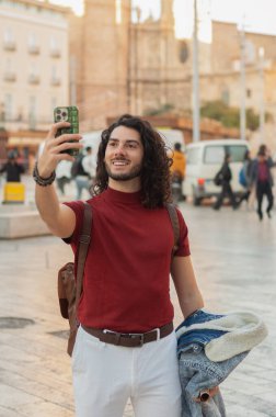 Uzun kıvırcık saçlı genç adam şehir meydanında akıllı telefonuyla bir selfie çeker ve seyahat macerasının özünü yakalar.