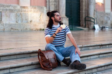 Uzun saçlı genç adam, güneşli bir günde Avrupa 'daki bir kilisenin önünde sokak yemeklerinden hoşlanıyor. Şehir yaşamını ve tarihi mimariyi gözler önüne seriyor.