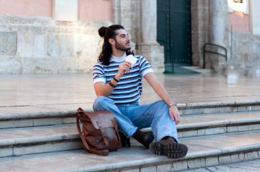 Uzun saçlı ve sakallı genç adam kilise merdivenlerinde komünyon ekmeği yiyor ve Avrupa 'daki bir şehre bakıyor. Günlük kıyafetleri ve deri çantası tefekkür sahnesine ekleniyor.
