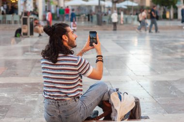 Uzun saçlı genç bir turist güneşli bir meydanda rahat bir şekilde oturarak canlı atmosferin tadını çıkarırken akıllı telefonuyla canlı anları yakalıyor.