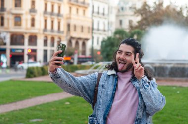 Uzun, kıvırcık saçlı genç adam canlı bir şehir parkında neşeli bir selfie çekiyor, dilini uzatıyor ve barış işareti yapıyor, gençlik kültürünü kucaklıyor.