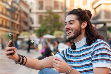 Uzun saçlı ve sakallı genç adam canlı bir şehir meydanında sosyal medyayı modern bir zamanda sokak hayatıyla harmanlarken akıllı telefonuyla mutlu bir selfie çekiyor.