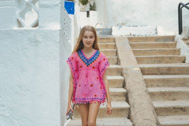 Canlı pembe bir tunik giyen genç bir kız güneşli, kıyı manzarasında resimli adımlar atıyor..