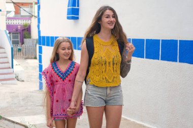 Genç bir anne ve kız el ele tutuşup, beyaz ve mavi duvarları olan bir turist köyünde, güneşli bir tatil gününün tadını çıkarıyorlar.