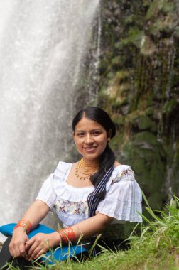 Geleneksel giysiler giymiş neşeli bir kadın bereketli doğanın ortasında huzurlu bir şelalenin yanında zarifçe poz verir.