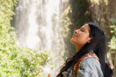 Genç bir kadın, yemyeşil ve doğal güzelliklerle çevrili çarpıcı bir şelalenin yanında sükunetin tadını çıkarır.