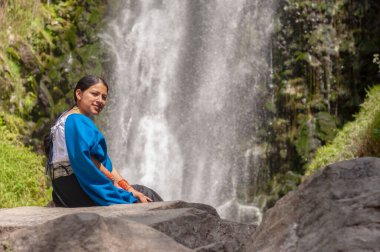 Bir kadın zarif ve sakin bir şelalenin yanında oturuyor. Etrafı yemyeşil bir manzara ve doğa ile çevrili.