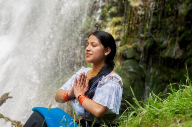 Genç bir kadın, güzel bir şelalede huzur içinde meditasyon yaparken, doğayı kucaklayarak, sesleri ve manzaraları sakinleştirir.