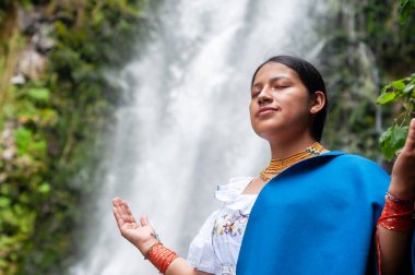 Geleneksel kıyafetlerle süslenmiş genç bir kadın çarpıcı bir şelalenin yanında doğanın huzur dolu özünü kucaklıyor.