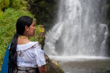 Huzurlu bir kadın görkemli ve nefes kesici bir şelalenin yanında durup zengin kültürel mirasını sergiliyor.