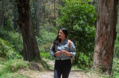 Bir kadın sakin bir orman yolunda yürüyor, tamamen yemyeşil ve uzun ağaçlarla çevrili.