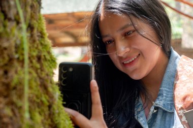 Genç bir kadın neşeli bir şekilde gülümser, akıllı telefonunu kullanarak, dingin, Mosscovered ağacının yanında.