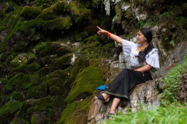 Yosun tutmuş bir kayanın üzerinde oturan bir kadın canlı yeşil bir ortamda doğayı işaret ederek sükuneti kucaklıyor..