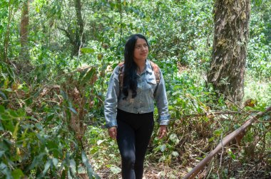 Bir kadın dolambaçlı bir orman patikasında güvenli adımlarla yürüyor, canlı yeşillikler ve yüksek ağaçlarla çevrili.