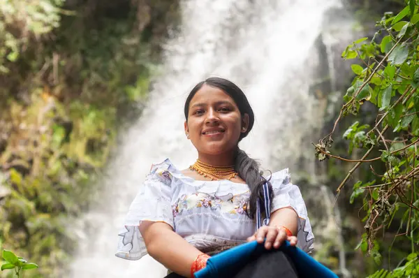 Güzel bir şelalenin kenarında gülümseyen genç bir kız, geleneksel giysiler içinde, doğayla çevrili..