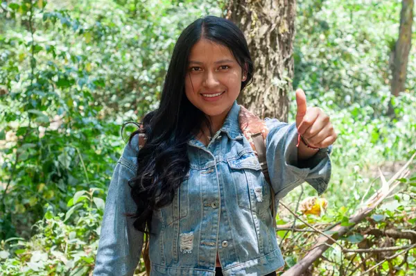 Neşeli genç bir kadın gülümseyip poz veriyor. Parlak başparmaklarıyla, yemyeşil bir ormanda.