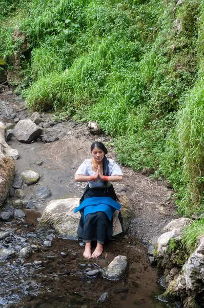 Genç bir kadın, yemyeşil ve canlı doğal güzelliklerle çevrili sakin bir derenin kenarında huzur içinde meditasyon yapıyor.