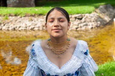 Huzurlu ve huzurlu bir portre. Huzurlu bir derenin yanında geleneksel kıyafetlerle süslenmiş genç bir kadını sergiliyor.