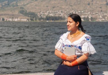 Geleneksel kıyafetli bir kadın dingin bir gölün kenarında zarif bir şekilde poz verir, canlı kültürel mirasını somutlaştırır.