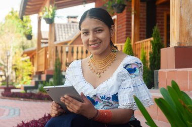 Geleneksel giyinmiş genç bir kadın çarpıcı bir açık hava ortamında modern bir tabletle meşgul.