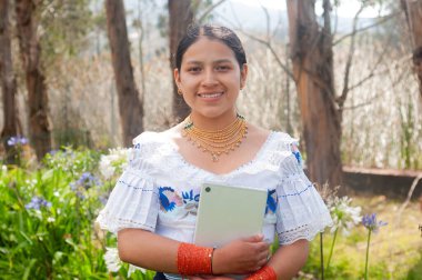 Geleneksel giysiler içinde genç bir yerli kadın, canlı çiçeklerin arasında bir tabletle gururla durur.