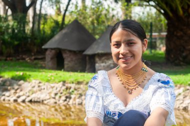 Geleneksel giysiler içinde genç bir yerli kadın yakınlardaki sakin bir suyun yanında zarifçe poz veriyor.