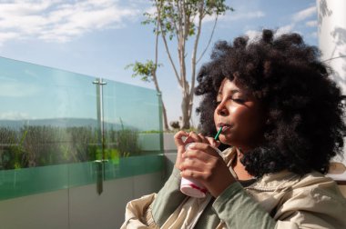 Genç bir kadın serinletici bir içkiyi yudumlarken yeşillik dolu güneşli bir manzaranın tadını çıkarıyor.