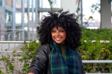 Neşeli bir kadın açık havada poz veriyor, doğal kıvırcık saçlarını ve şık eşarbını modern bir arka planda sergiliyor..