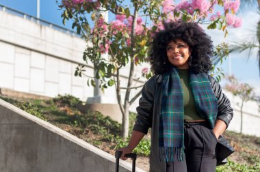 Canlı bir kadın dışarıda duruyor, ışıl ışıl gülümsüyor ve kendine güvenini ve tarzını güzel bir ortamda sergiliyor.