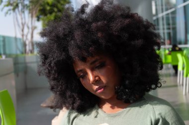 Şehvetli bir afro saç stili olan düşünceli genç bir kadın modern bir açık havada rahatça oturuyor.