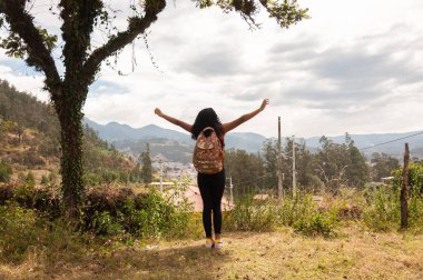 Sırt çantası takan genç bir kadın, mutluluk içinde kollarını açmış, dışarıda huzur içinde eğleniyor.