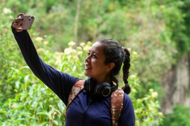 Neşeyle dolu genç bir kadın yemyeşil ve manzaralı bir yerin ortasında canlı bir selfie çeker.