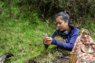 Genç bir kadın yemyeşil bir ortamda mutlu bir şekilde oturur, güzel doğayla çevrili telefonuna odaklanır.
