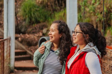 İki neşeli genç Latin Amerikalı kadın bir köprüde duruyor, manzaranın tadını çıkarıyorlar, doğanın ve arkadaşlığın güzelliğini kucaklarken gülücüklerini ve kahkahalarını paylaşıyorlar.