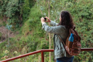 Sırt çantası ve su şişesiyle gezen kadın turist akıllı telefonuyla güzel yeşil bir manzaranın fotoğraflarını çekiyor ve doğada rahatlatıcı bir günün tadını çıkarıyor.