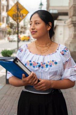 Geleneksel giysiler içinde genç bir kadın, bir şehir caddesinde elinde dosyaları tutarak güvenle duruyor ve geleceğe bakıyor..