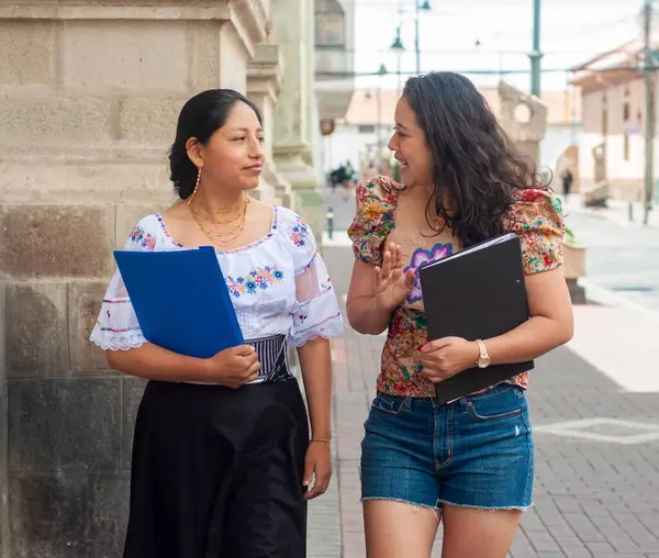 İki kadın, biri geleneksel elbiseli, diğeri ise modern, hayat dolu canlı bir şehir ortamında sohbet ediyorlar.