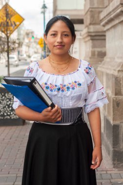 Şehir ortamında, renkli geleneksel kıyafetler giymiş bir kadın gururla bir cilt tutar.