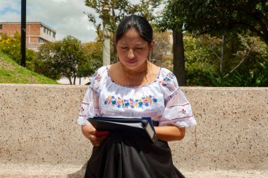 Zarif bir şekilde geleneksel kıyafetlerini giymiş bir kadın huzurlu bir açık hava ortamında tablete odaklanıyor.