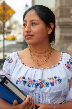 Geleneksel giyinmiş gururlu bir kadın, elinde kültürel kimlik ve eğitimi simgeleyen bir ciltle dışarıda duruyor.