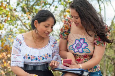 Geleneksel kıyafetlerle süslenmiş iki kadın gezici bir cihazla uğraşırken açık havada eğleniyorlar.