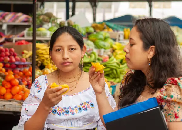 İki kadın rengarenk ürünlerle dolu hareketli, canlı bir pazarda taze, olgun meyveleri tadıyor.