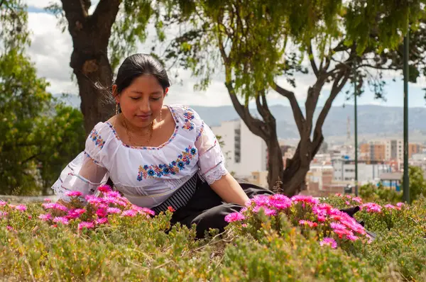 Geleneksel giysiler içinde zarif giyinmiş bir kadın çarpıcı bir açık hava ortamında çeşitli canlı çiçeklere hayran kalır.