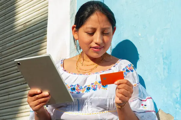 Canlı ve renkli bir ortamda, geleneksel giyinmiş bir kadın tabletini ve kredi kartını kullanır.