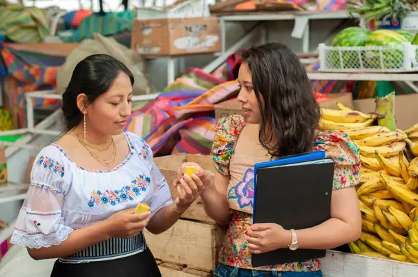 Canlı, geleneksel giyinmiş iki kadın canlı bir pazarın ortasındaki taze meyveleri dikkatle inceliyor.
