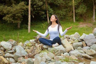 Huzurlu bir kadın, güzel bir doğal ortamda huzur içinde meditasyon yapar. Etrafı kayalar ve yemyeşil ağaçlarla çevrilidir.