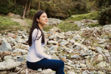 Genç bir kadın, sakin bir doğal ortamda kayaların arasında huzur içinde oturur ve yalnızlığını tamamen benimser.