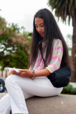 Genç bir kız büyüleyici bir kitaba dalmış, açık havada, yemyeşil ve güzel bir park alanında oturuyor.